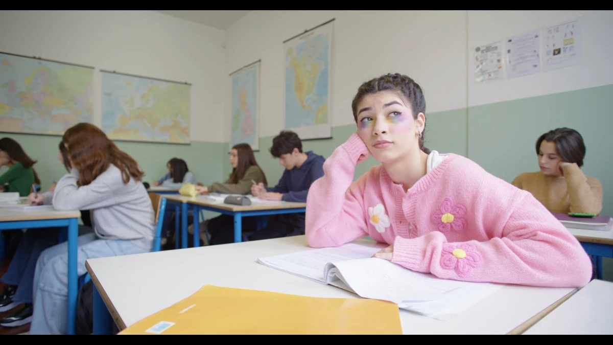 Bright source frame from Leumos demo footage: a classroom, a student in a pink cardigan resting her chin on her hand, white and pale green walls, wall maps behind